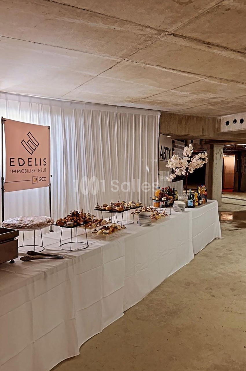 Table dressée avec des amuse-bouches, nappée de blanc, dans un espace en béton avec une bannière ’Edelis Immobilier Neuf’.