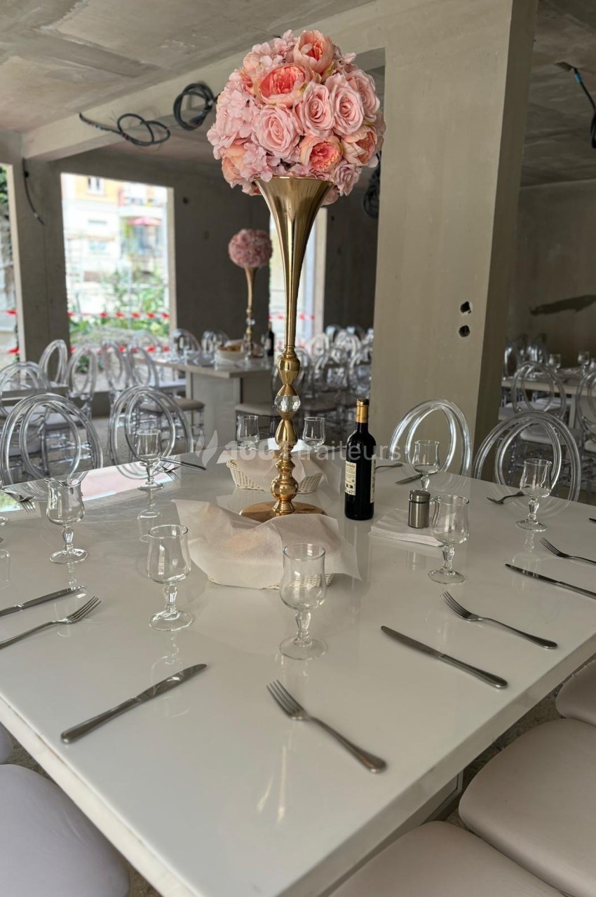 Table dressée avec nappes blanches, couverts, verres, centre de table floral rose et bouteille de vin dans une salle moderne.