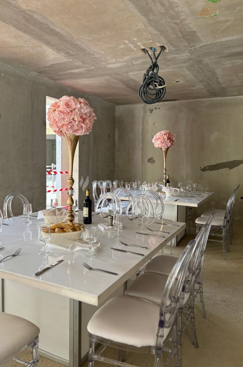 Salle avec tables dressées, chaises transparentes, centres de table fleuris roses et murs en béton brut.