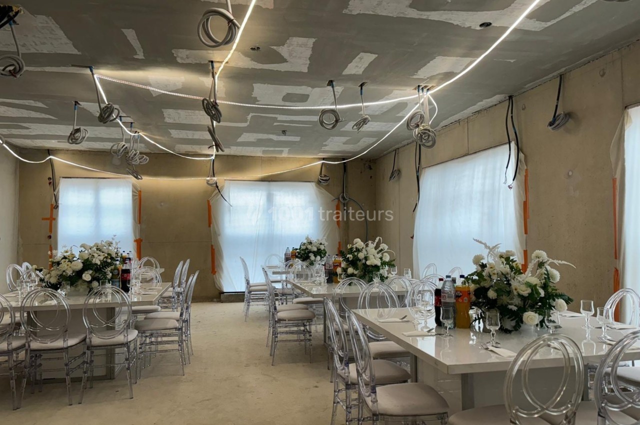 Salle de réception en cours d'aménagement, avec tables décorées de fleurs blanches et chaises transparentes.