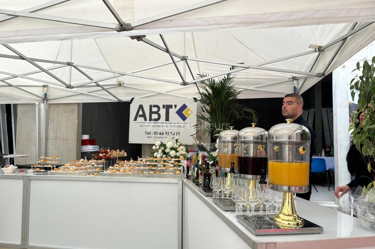 Buffet sous une tente blanche avec boissons en distributeurs et amuse-bouches, un homme en arrière-plan.