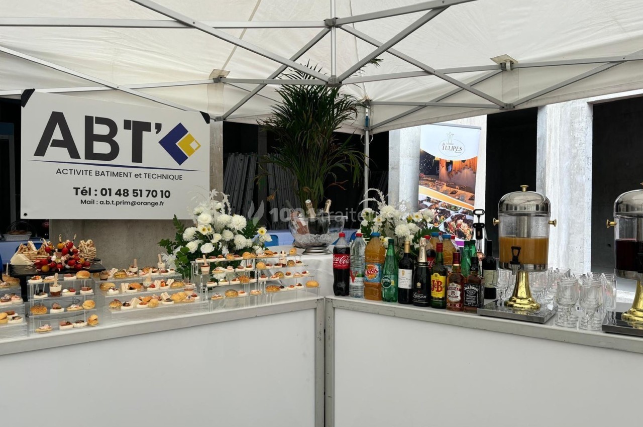 Buffet sous une tente avec boissons, amuse-bouches et décoration florale, devant un panneau d'entreprise ABT.