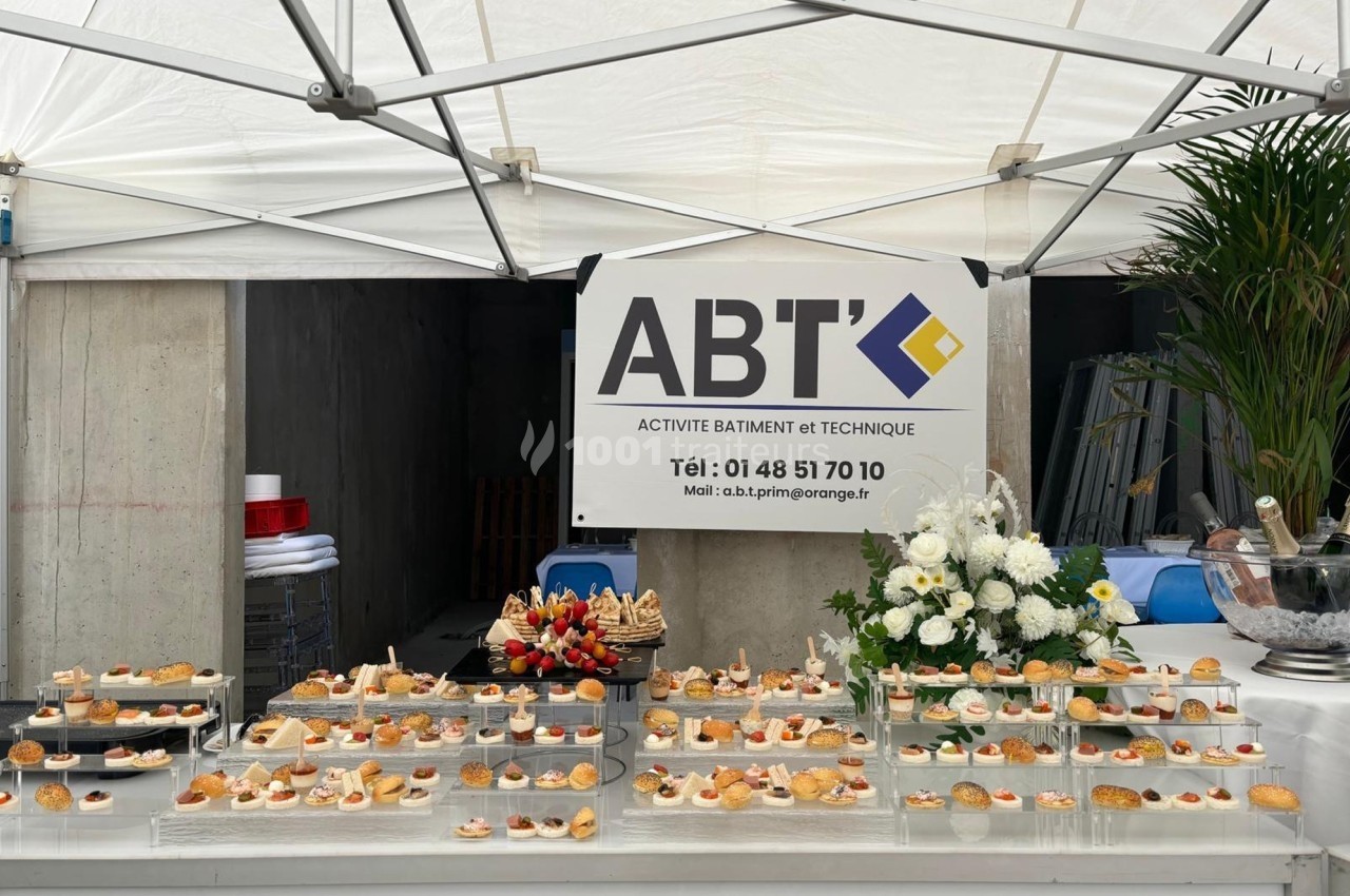 Buffet de petits fours disposé sous une tente blanche, avec un panneau d'entreprise en arrière-plan.