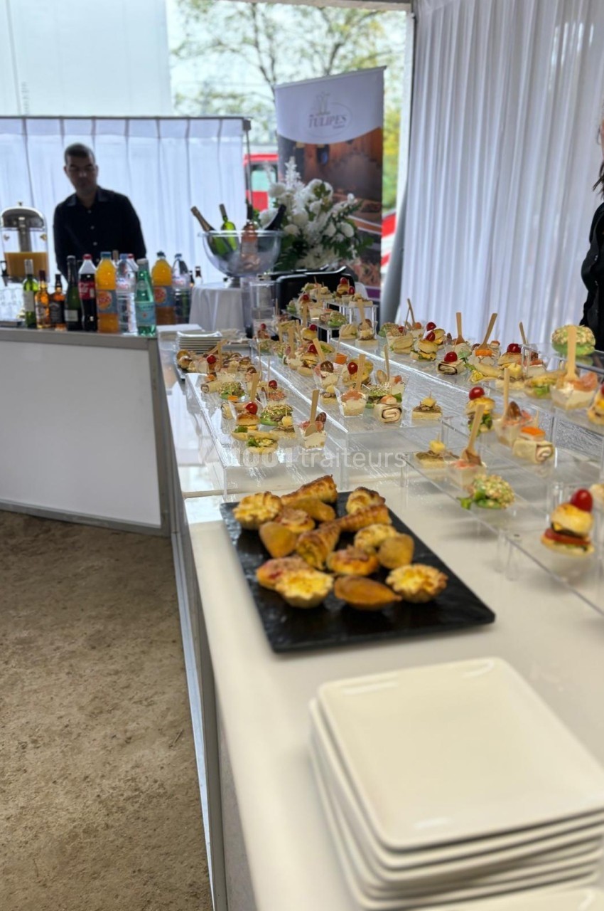 Buffet de cocktails avec amuse-bouches variés disposés sur une table blanche, boissons et personnel en arrière-plan.