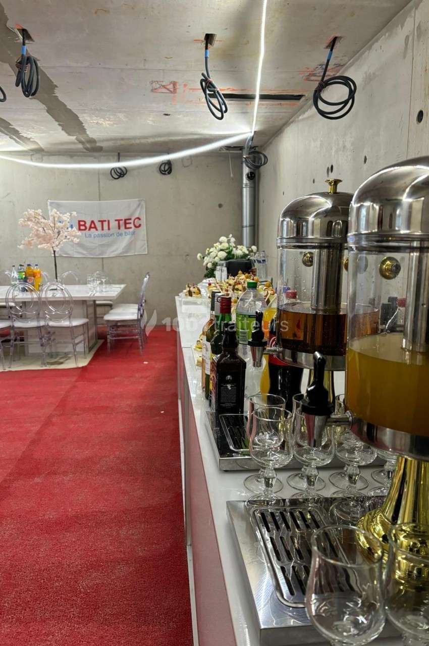 Buffet avec boissons et verres sur une table, dans une salle au sol rouge et murs en béton brut.