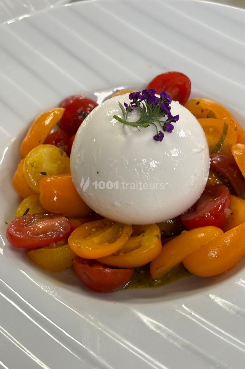 Assiette de burrata garnie de tomates cerises colorées et décorée d'une fleur violette sur une assiette blanche.