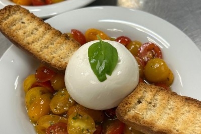 Assiette de burrata garnie de tomates cerises colorées et décorée d'une fleur violette sur une table blanche.