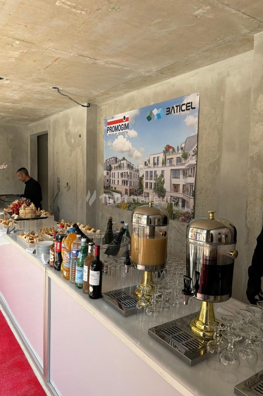 Buffet avec boissons, viennoiseries et fruits, installé devant des affiches promotionnelles de projets immobiliers.