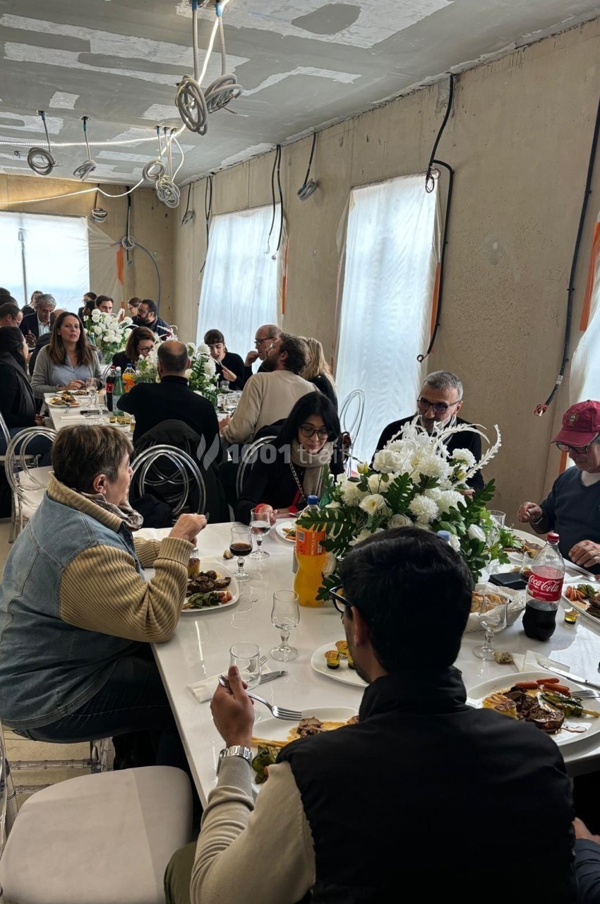 Des personnes partagent un repas autour de longues tables décorées de fleurs dans une salle lumineuse.
