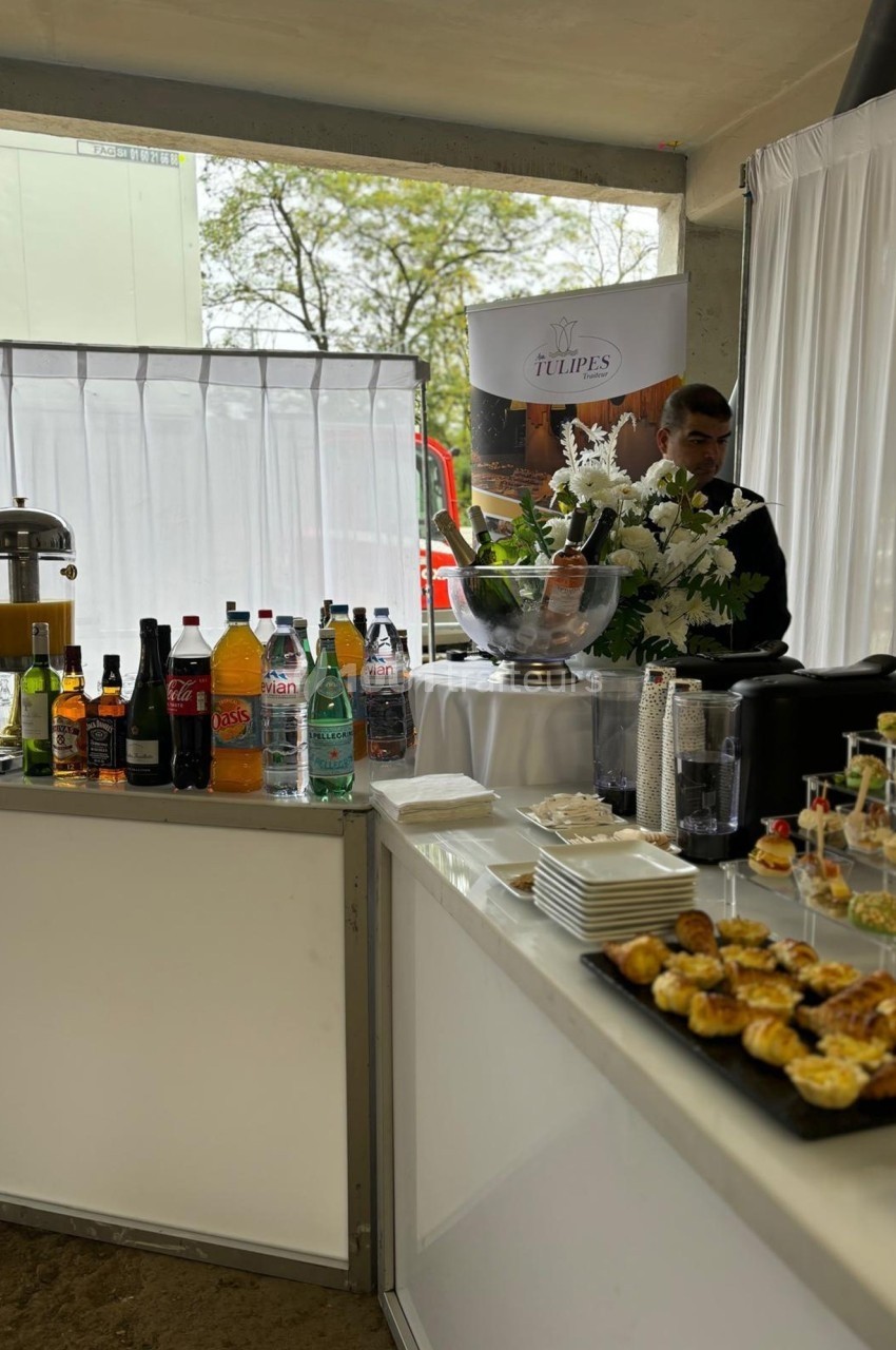 Buffet avec boissons variées, viennoiseries et fleurs blanches, disposé dans un espace lumineux avec rideaux blancs.