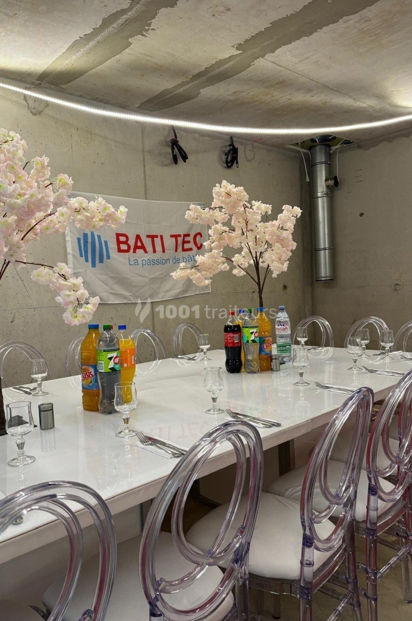 Table dressée avec des chaises transparentes, décorée de fleurs artificielles, boissons variées et fond avec mur brut.