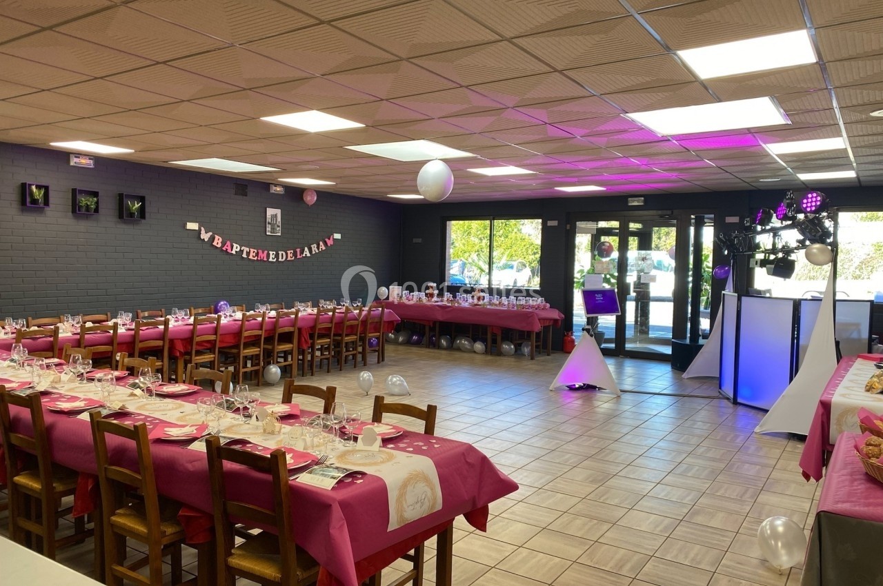Salle décorée pour une réception avec tables dressées en nappes roses, guirlande murale et espace DJ éclairé.