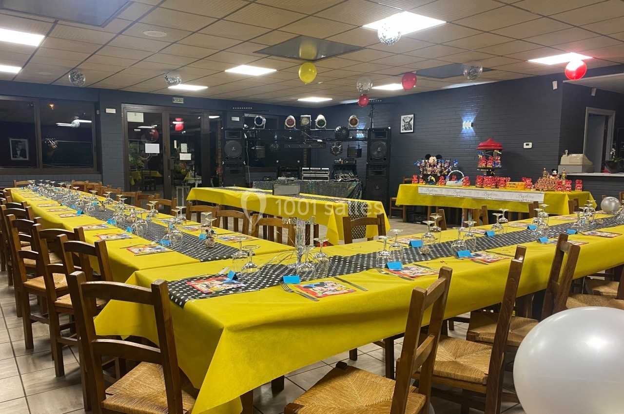 Salle décorée pour une fête avec de longues tables jaunes, chaises en bois et bonbonnières en arrière-plan.