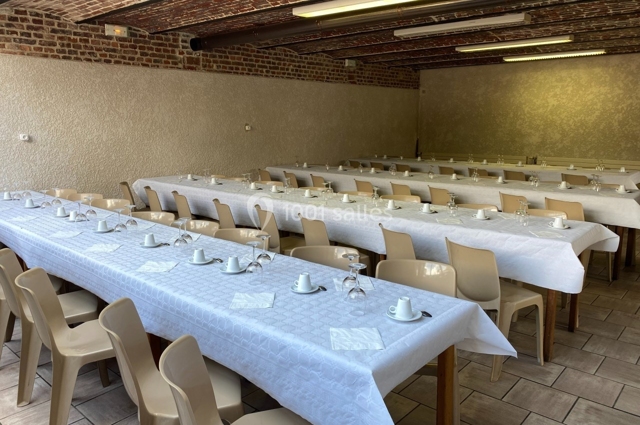 Salle avec de longues tables recouvertes de nappes blanches, disposées avec tasses, verres et serviettes.