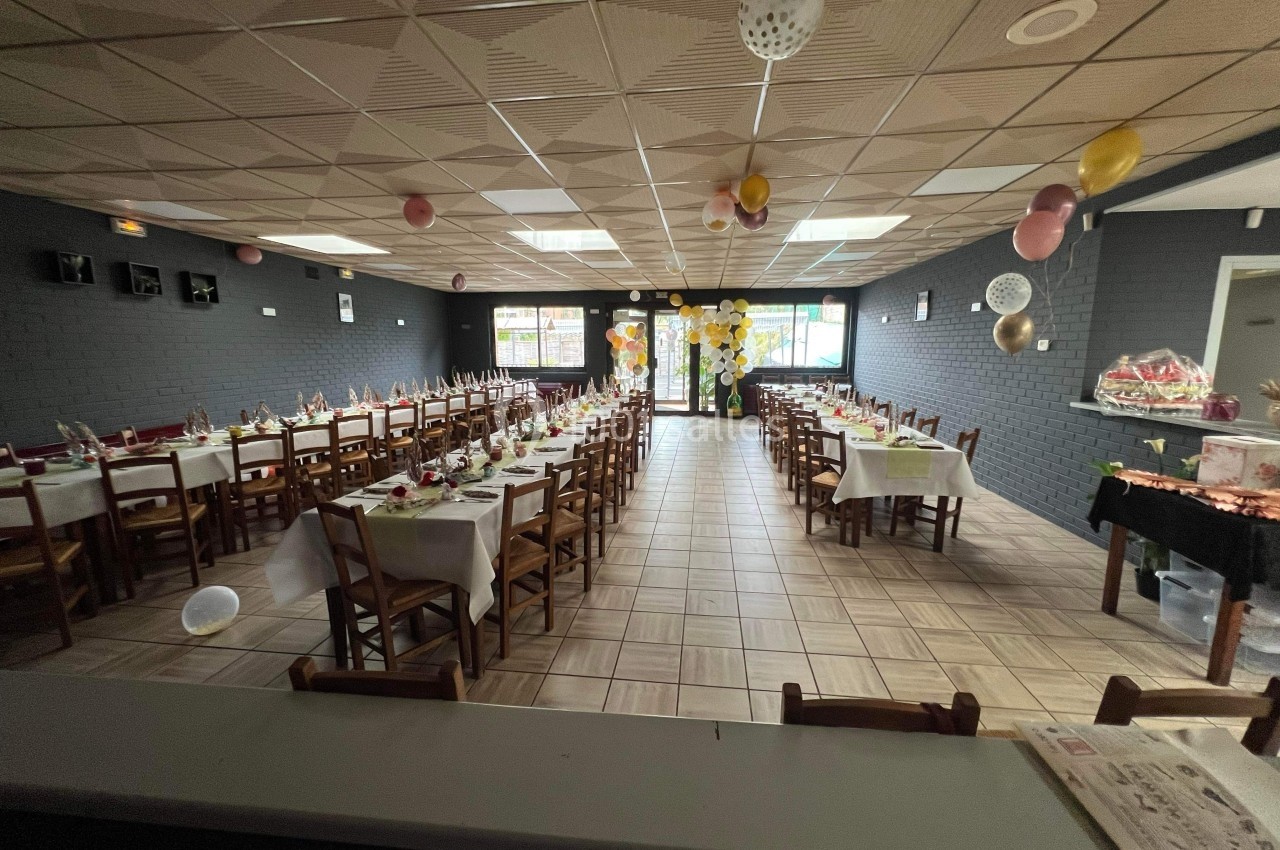 Salle décorée pour un événement, avec de longues tables dressées, des chaises en bois et des ballons suspendus.
