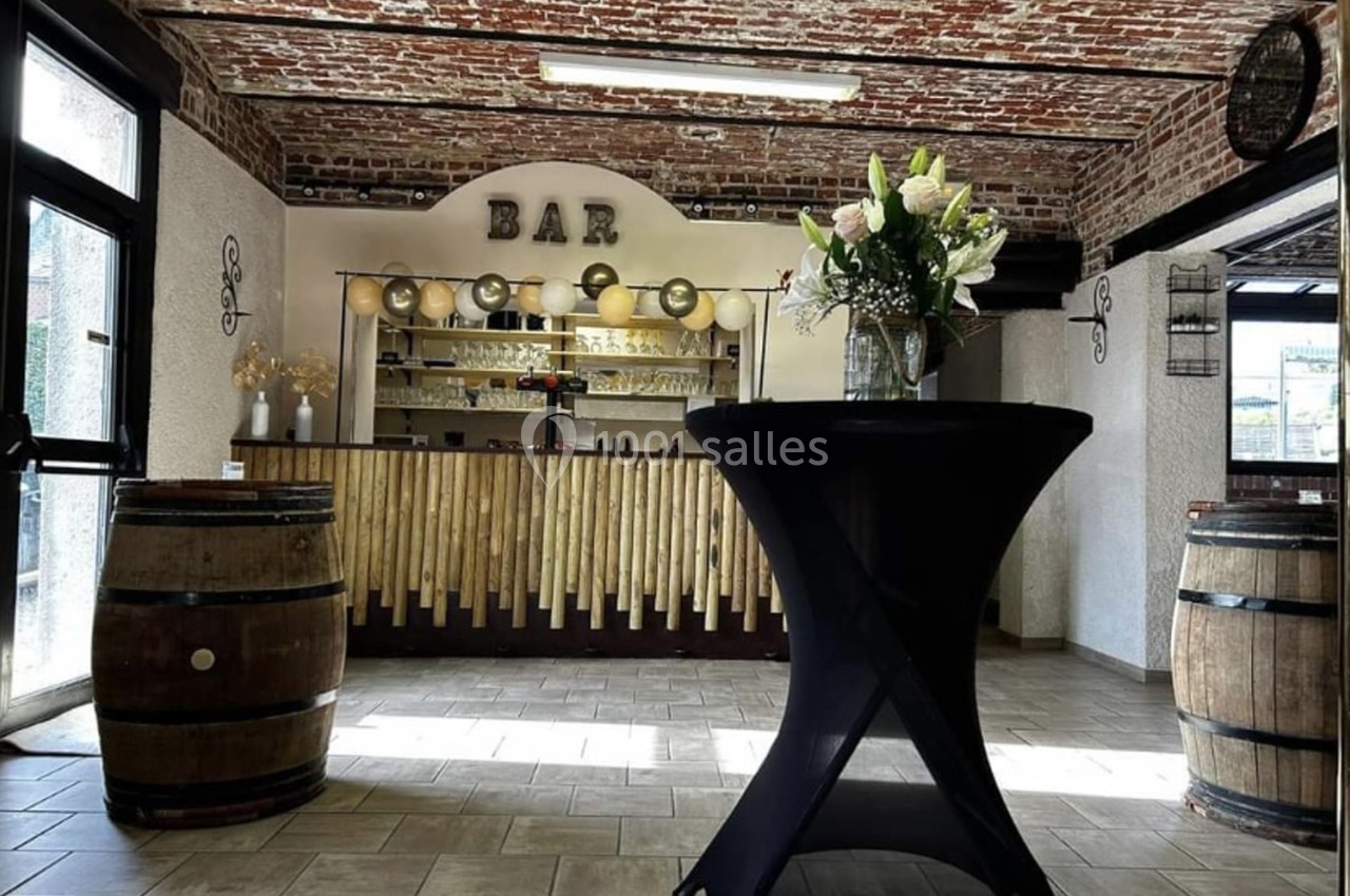 Salle avec un bar en bois, décorée de briques apparentes, tonneaux et une table haute ornée de fleurs blanches.