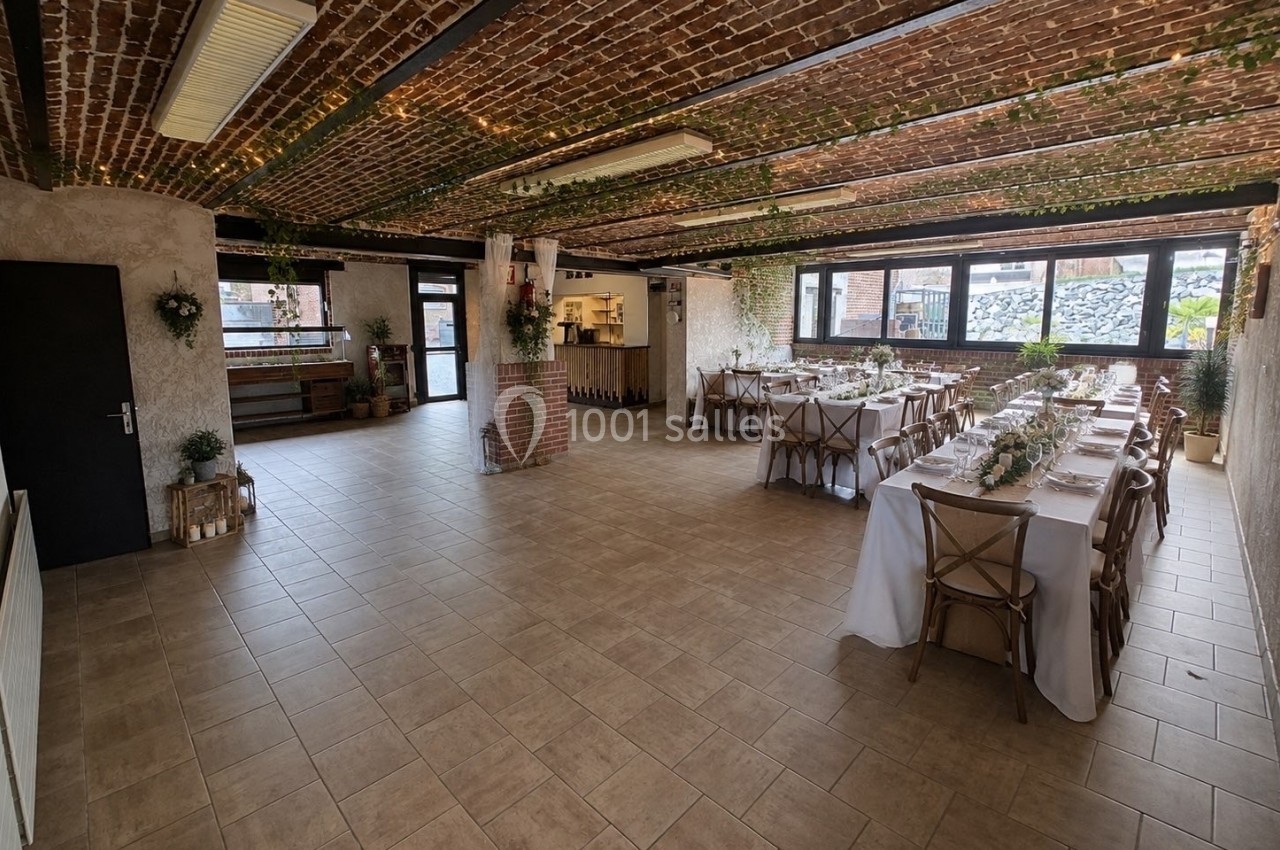 Salle de réception avec tables dressées, murs en briques, poutres décorées de verdure et lumière naturelle.