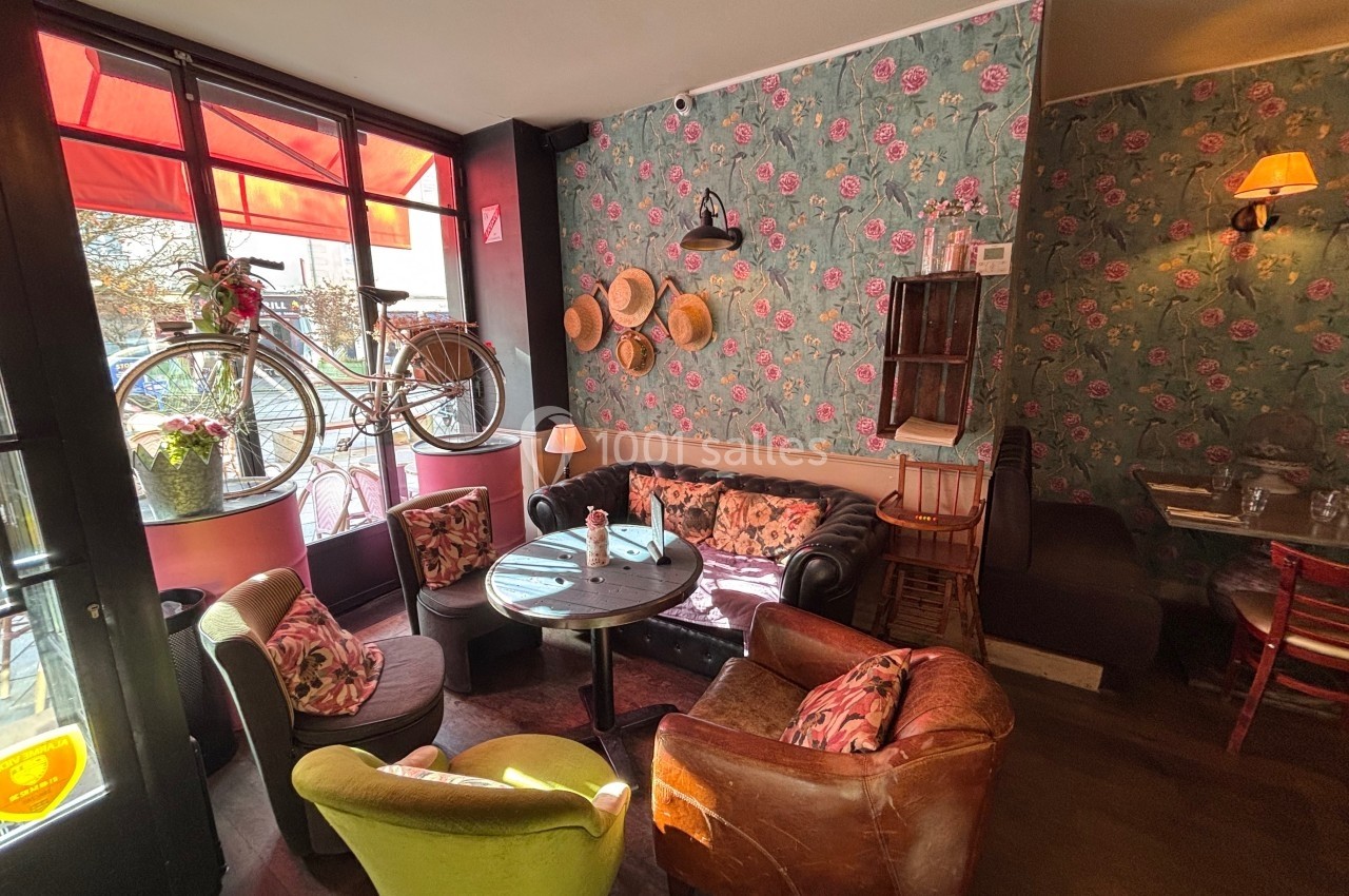 Salon de thé cosy avec fauteuils colorés, décoration florale et un vélo suspendu près d'une fenêtre lumineuse.
