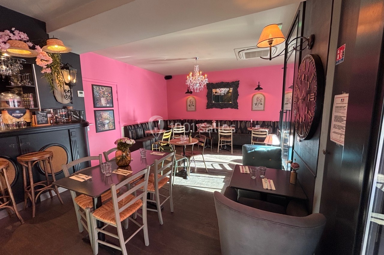 Salle de restaurant lumineuse avec murs roses, mobilier en bois et décoration élégante, incluant lustres et horloge murale.