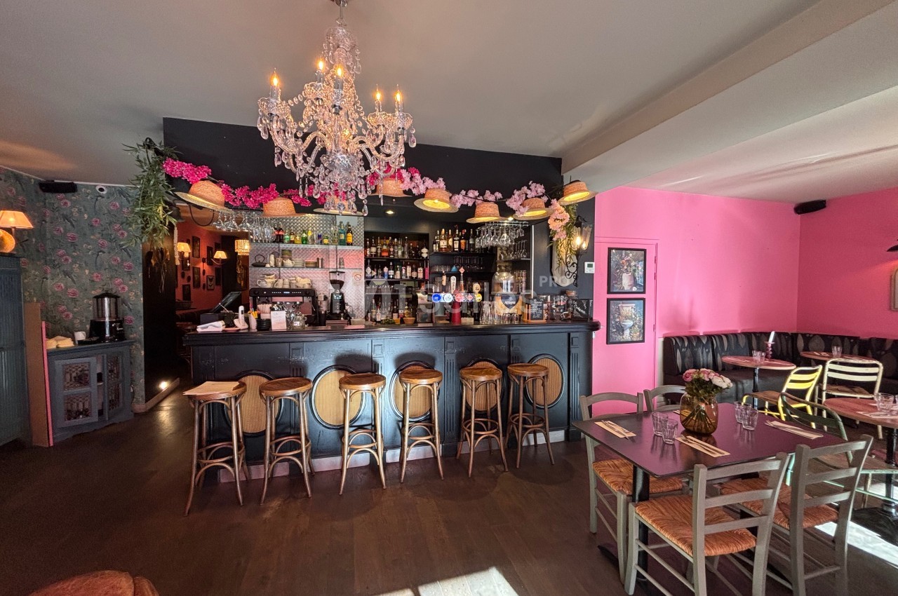 Intérieur d'un bar-restaurant avec comptoir noir, tabourets en bois, lustre et décoration florale colorée.