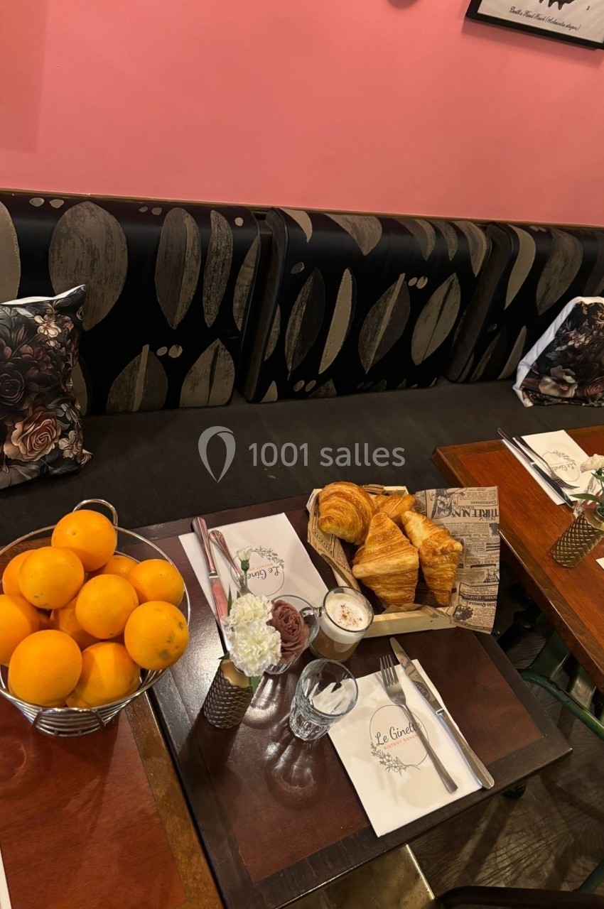 Table de petit-déjeuner avec croissants, confiture, oranges dans un panier et décoration florale sur une banquette.