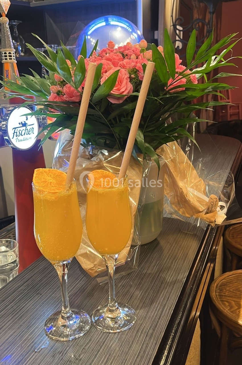 Deux verres de cocktail orange avec des pailles en papier, posés sur un comptoir devant un bouquet de fleurs roses.