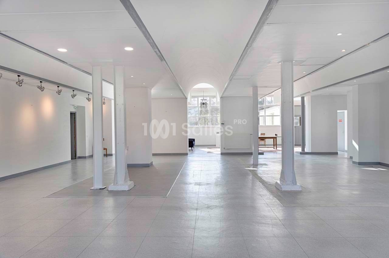Salle lumineuse et spacieuse avec des colonnes blanches, un sol en carrelage gris et de grandes fenêtres laissant entrer la…