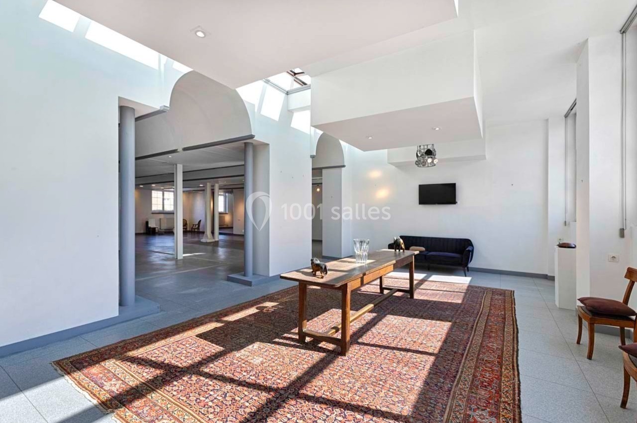 Salle lumineuse avec grandes fenêtres, tapis au sol, table en bois, canapé noir et murs blancs.