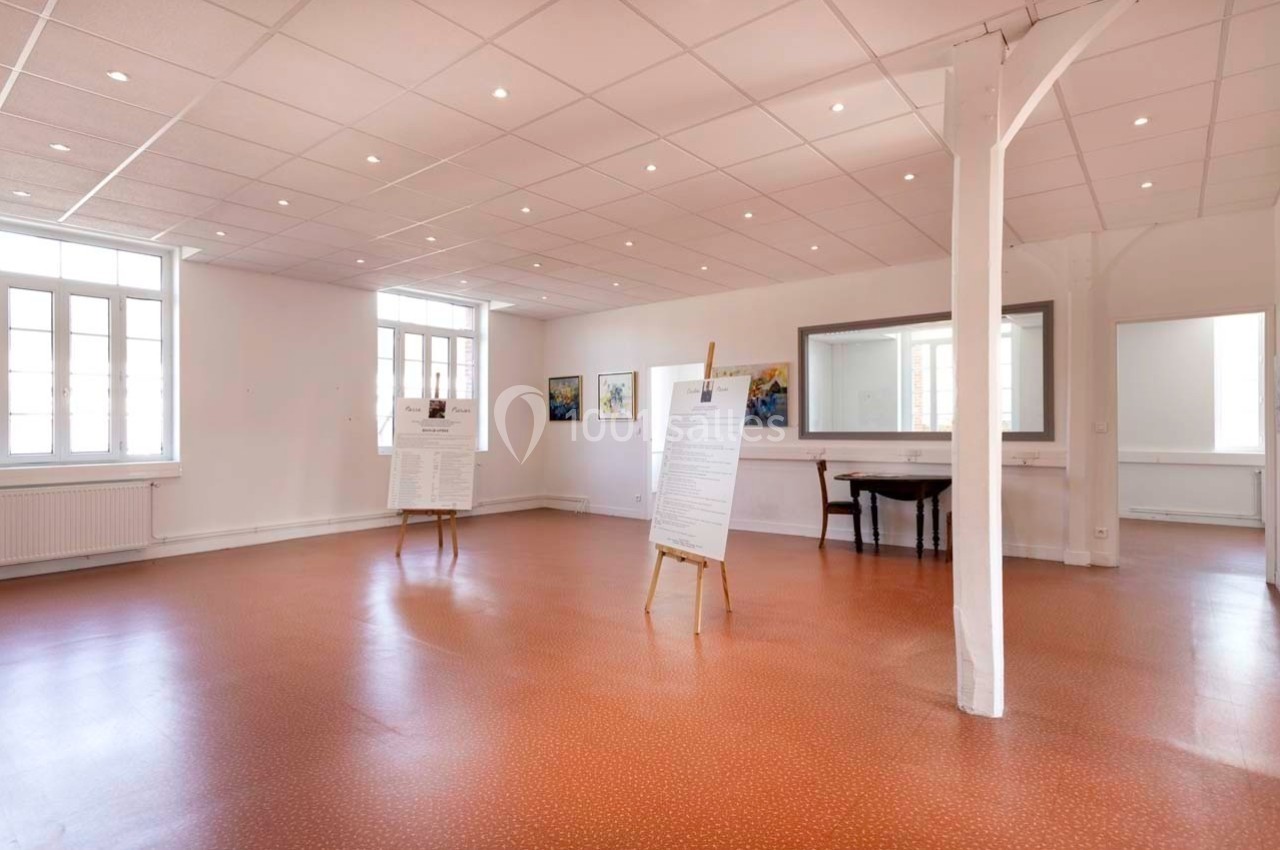 Salle lumineuse avec sol en carrelage orange, affiches sur chevalets et tableaux accrochés aux murs blancs.