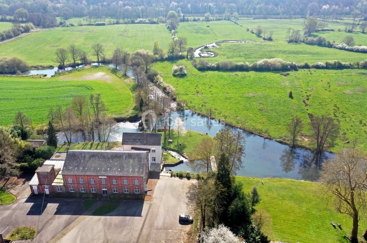 Vue aérienne d'un bâtiment en briques rouges entouré de prairies verdoyantes, d'arbres et de cours d'eau.