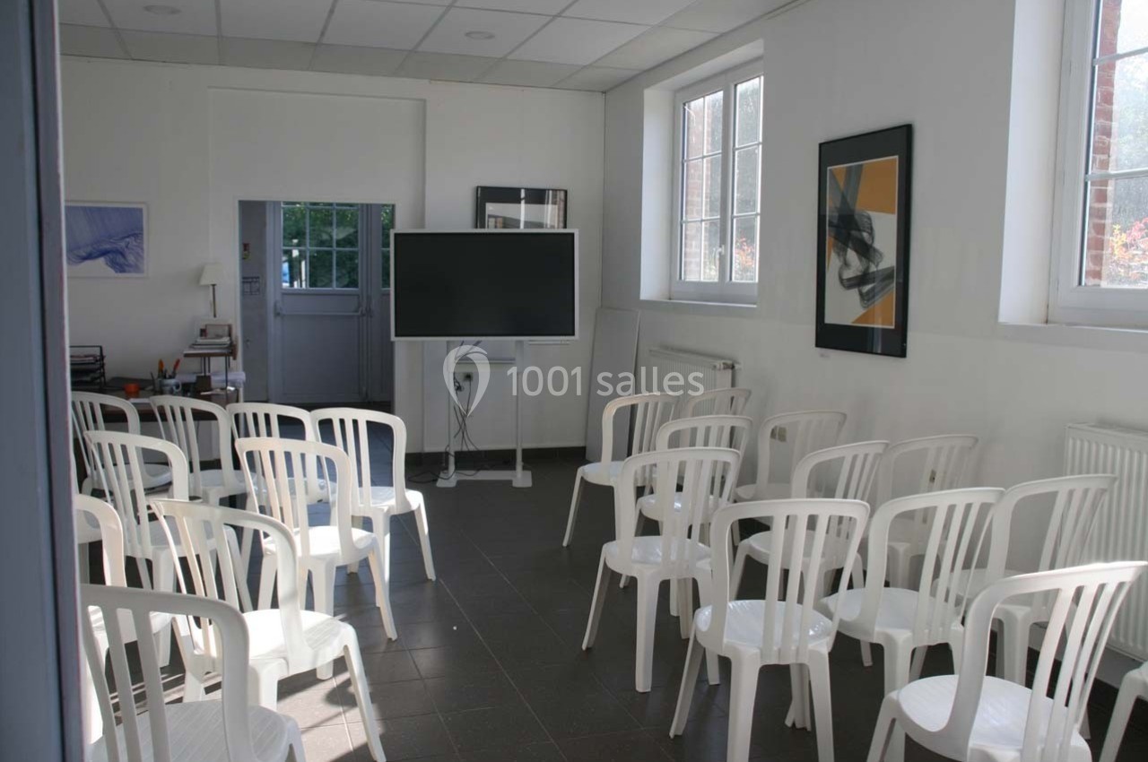 Salle lumineuse avec des chaises blanches disposées face à un écran noir sur pied, près de fenêtres et de tableaux.