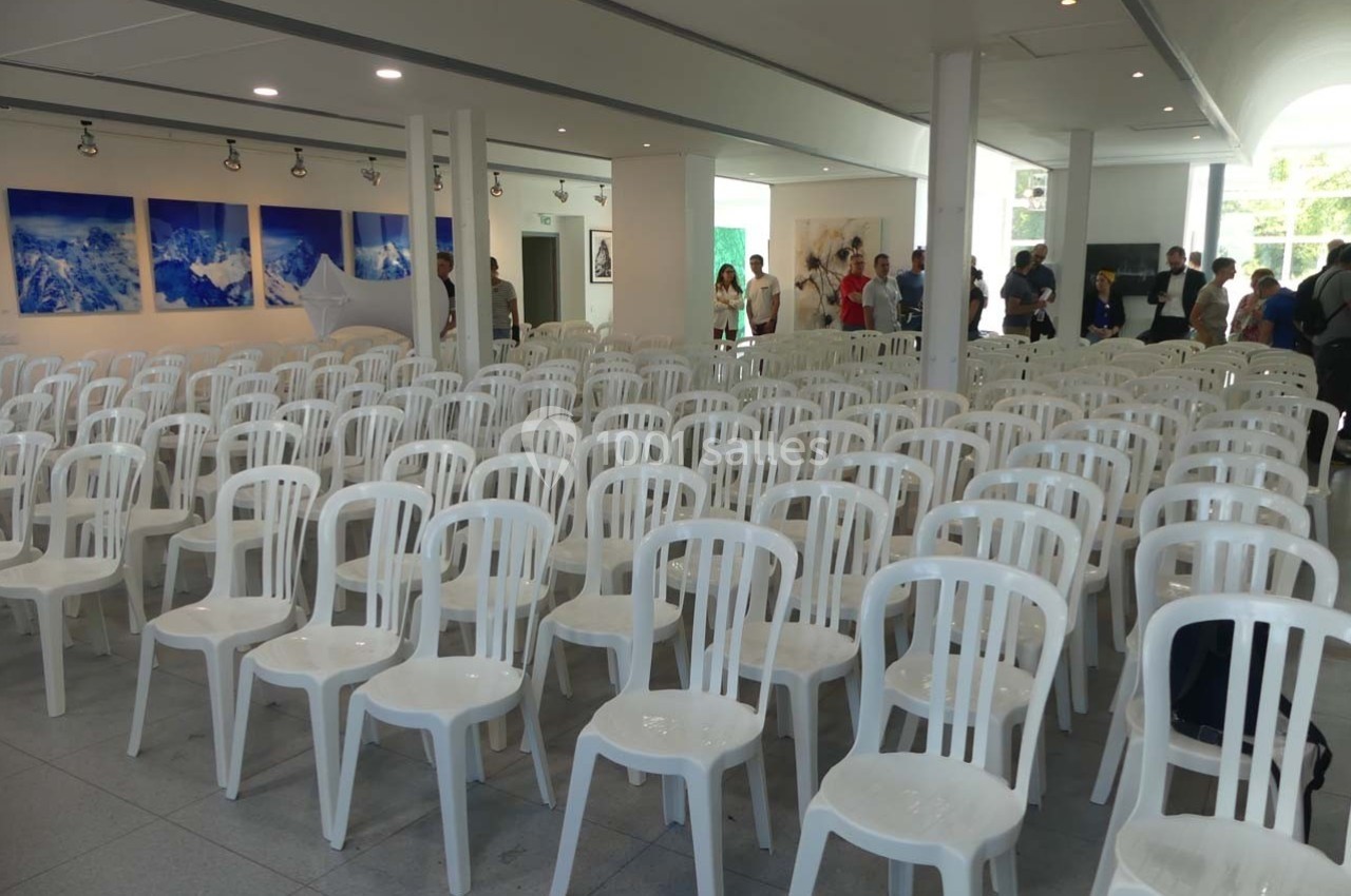 Salle lumineuse avec de nombreuses chaises blanches en plastique alignées, quelques personnes en arrière-plan.