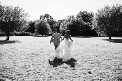 Miniature John Croizé Photographie #2 Un couple en tenue de mariage, l'homme portant la femme dans ses bras devant un mur en pierre ancienne.