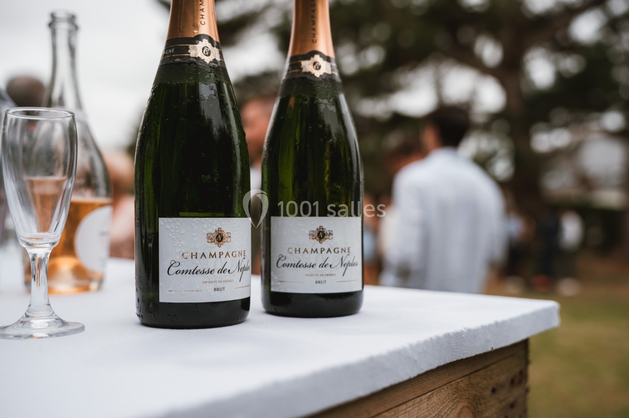 Deux bouteilles de champagne posées sur une table blanche, avec des verres et un arrière-plan flou de personnes.