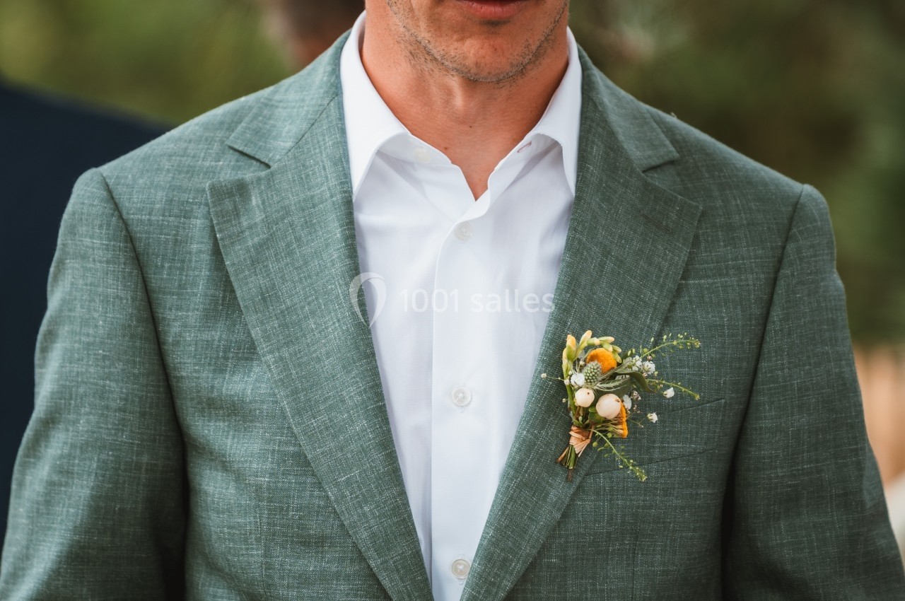 Homme en costume vert avec une boutonnière composée de fleurs et de baies colorées.