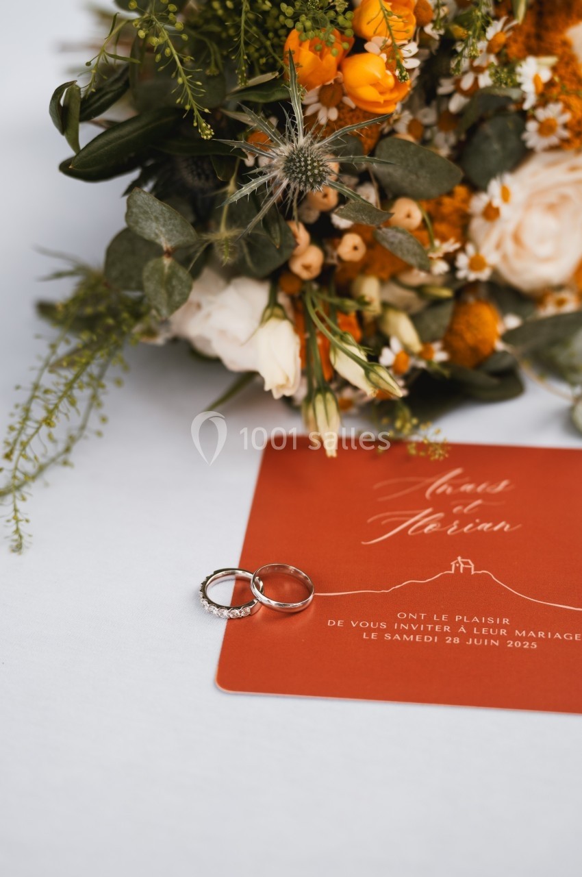 Deux alliances posées sur une carte d'invitation rouge, avec un bouquet de fleurs blanches et orangées en arrière-plan.