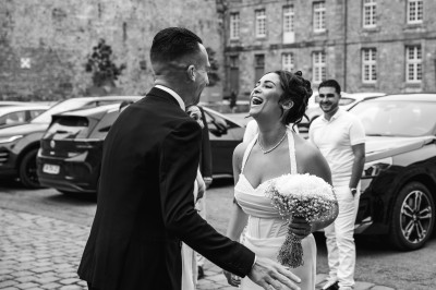 Miniature John Croizé Photographie #4 Un couple en tenue de mariage, l'homme portant la femme dans ses bras devant un mur en pierre ancienne.