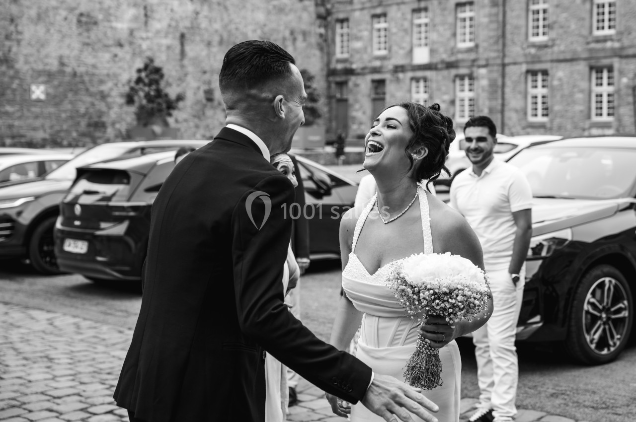 Un couple souriant en tenue de mariage se retrouve devant des voitures garées et un bâtiment en arrière-plan.