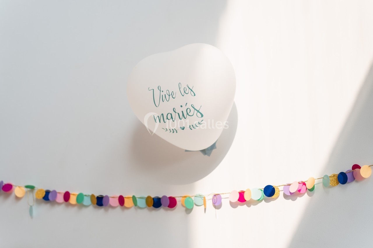 Ballon blanc avec l'inscription ’Vive les mariés’ suspendu au-dessus d'une guirlande colorée sur fond clair.