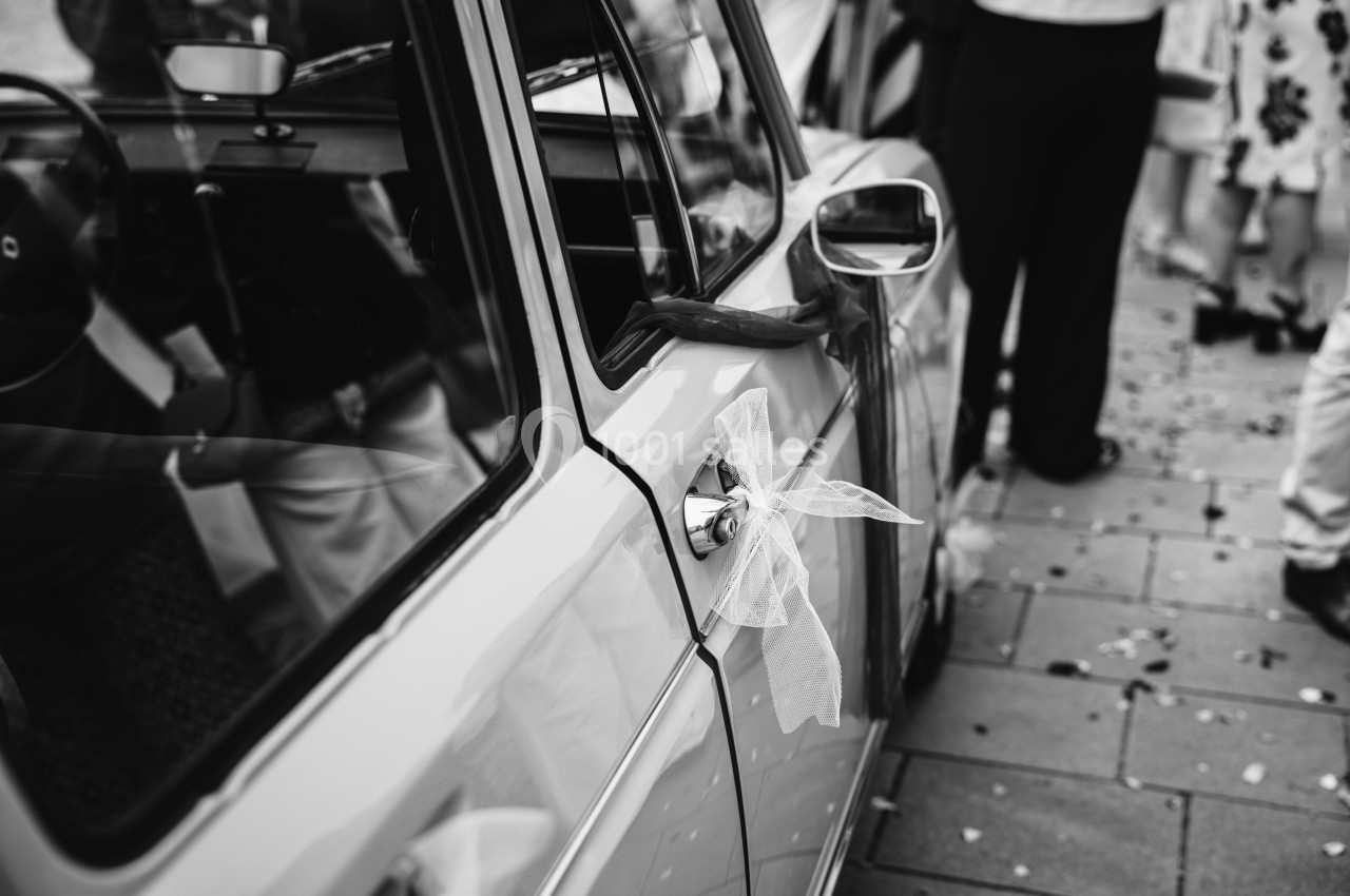 Ruban décoratif attaché à la poignée d'une voiture ancienne, entourée de personnes sur un trottoir pavé.