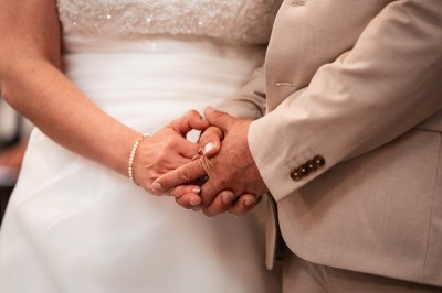 Miniature John Croizé Photographie #12 Un couple en tenue de mariage, l'homme portant la femme dans ses bras devant un mur en pierre ancienne.