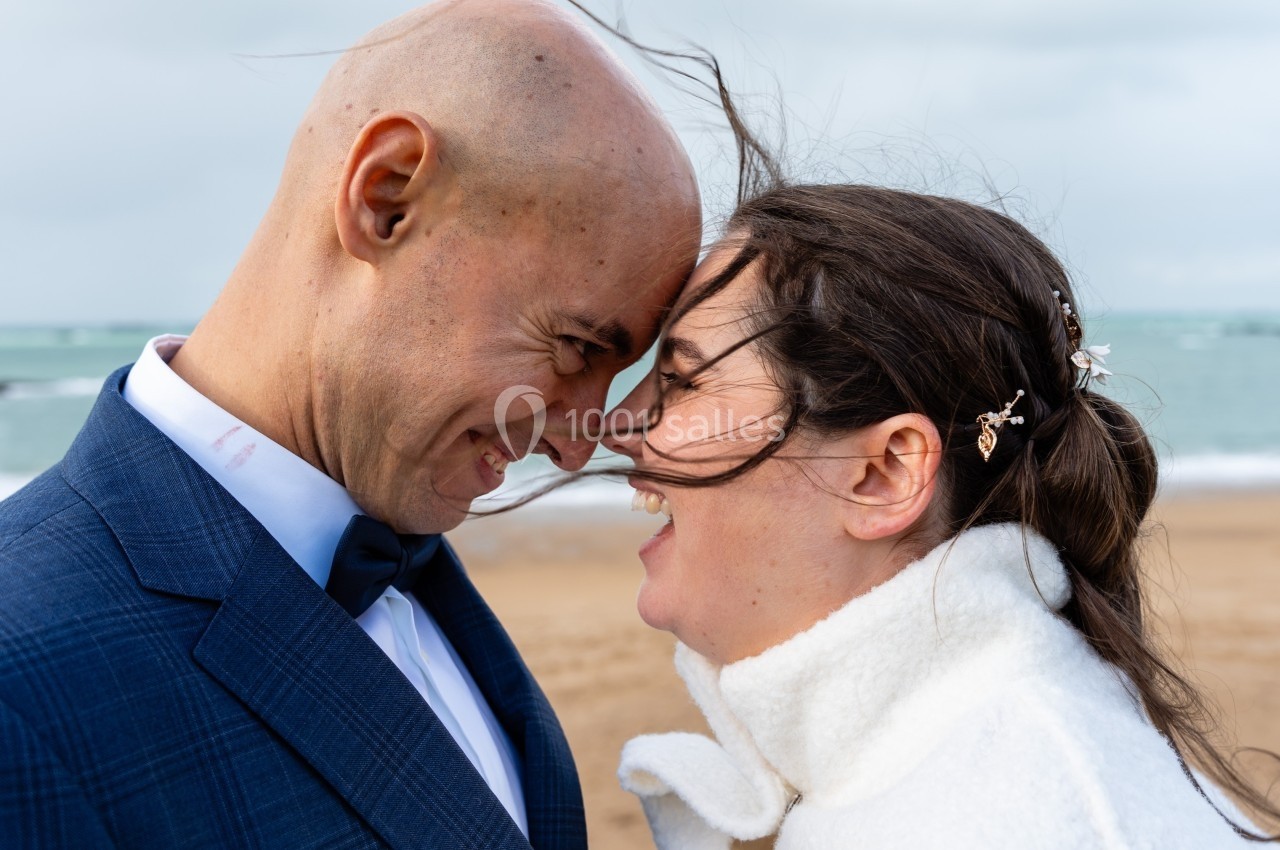 Un couple souriant face à face sur une plage venteuse, cheveux ébouriffés par le vent.