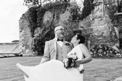 Miniature John Croizé Photographie #16 Un couple en tenue de mariage, l'homme portant la femme dans ses bras devant un mur en pierre ancienne.