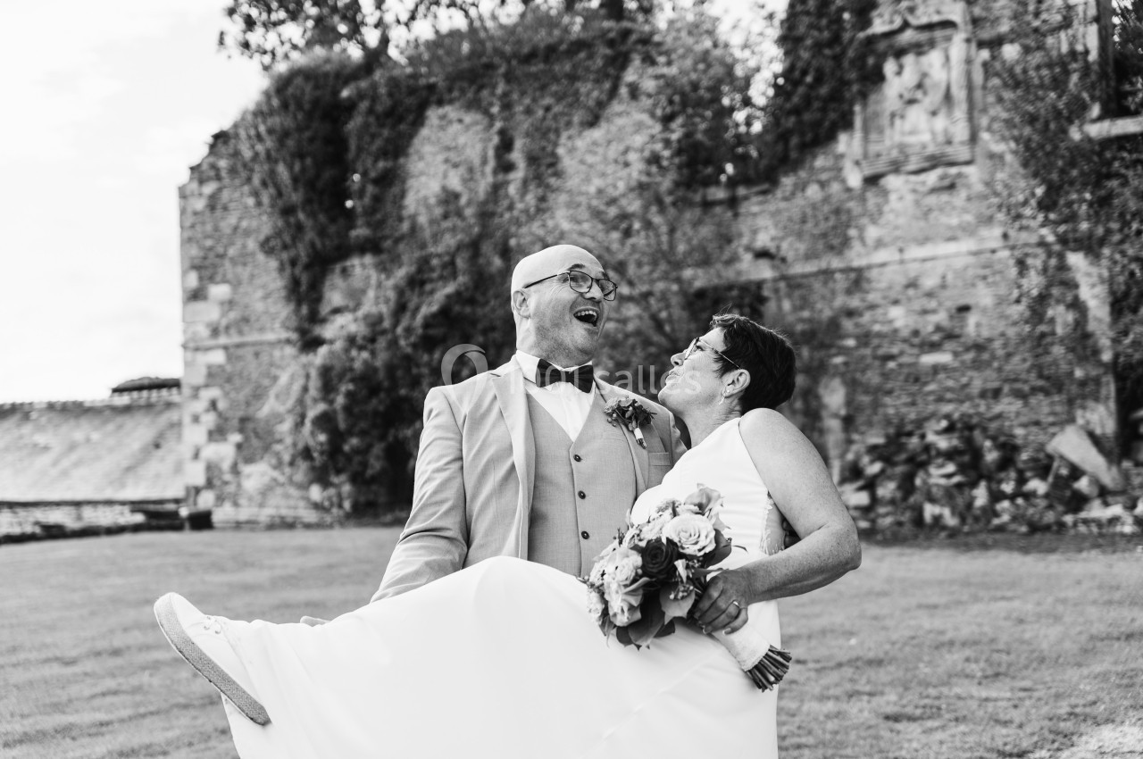 Un couple en tenue de mariage, l'homme portant la femme dans ses bras devant un mur en pierre ancienne.