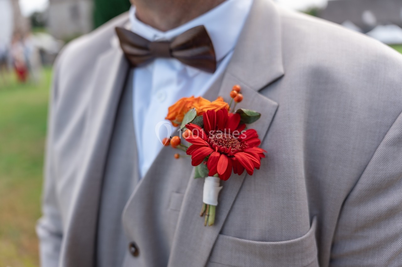 Homme en costume beige portant un nœud papillon marron et une boutonnière avec des fleurs rouges et orange.