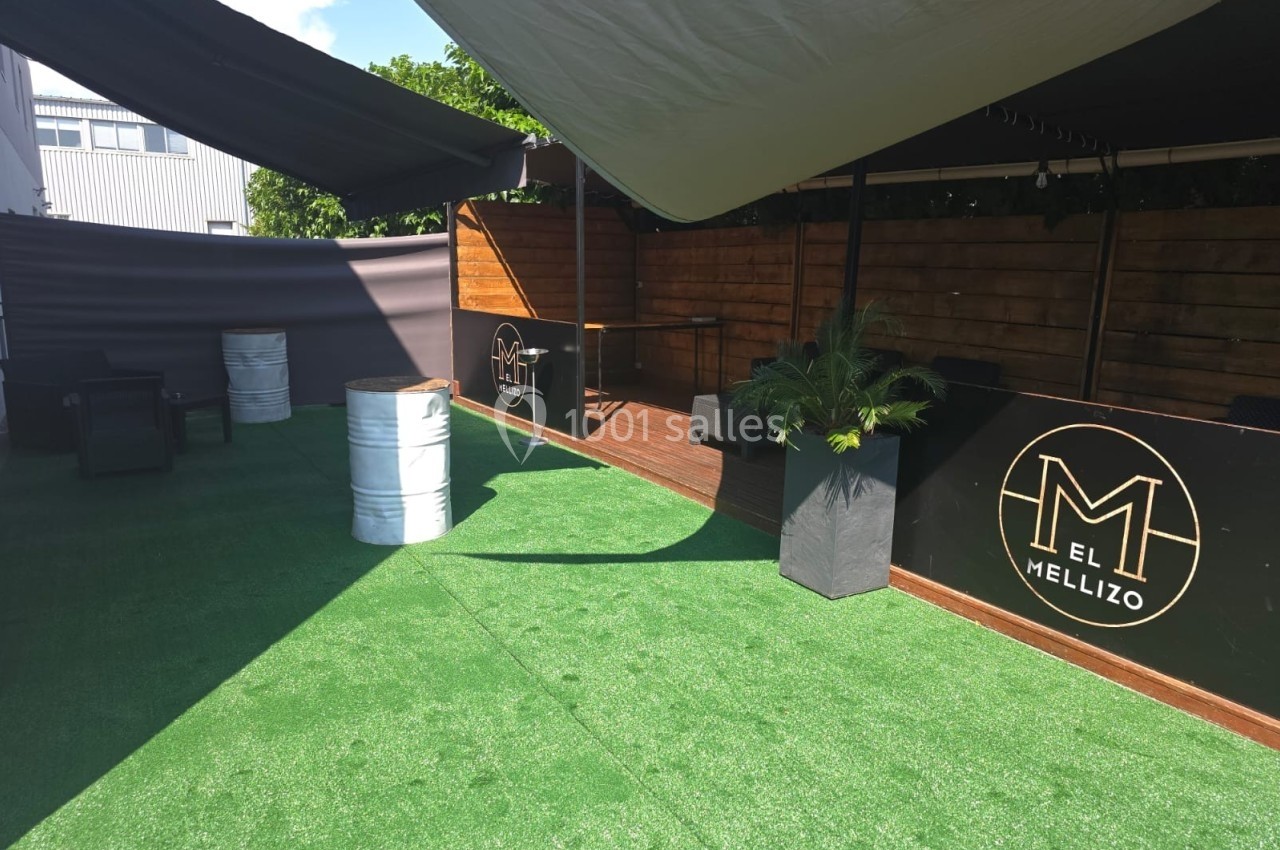 Espace extérieur avec sol en gazon synthétique, mobilier minimaliste et enseigne ’El Mellizo’ sur des panneaux noirs.