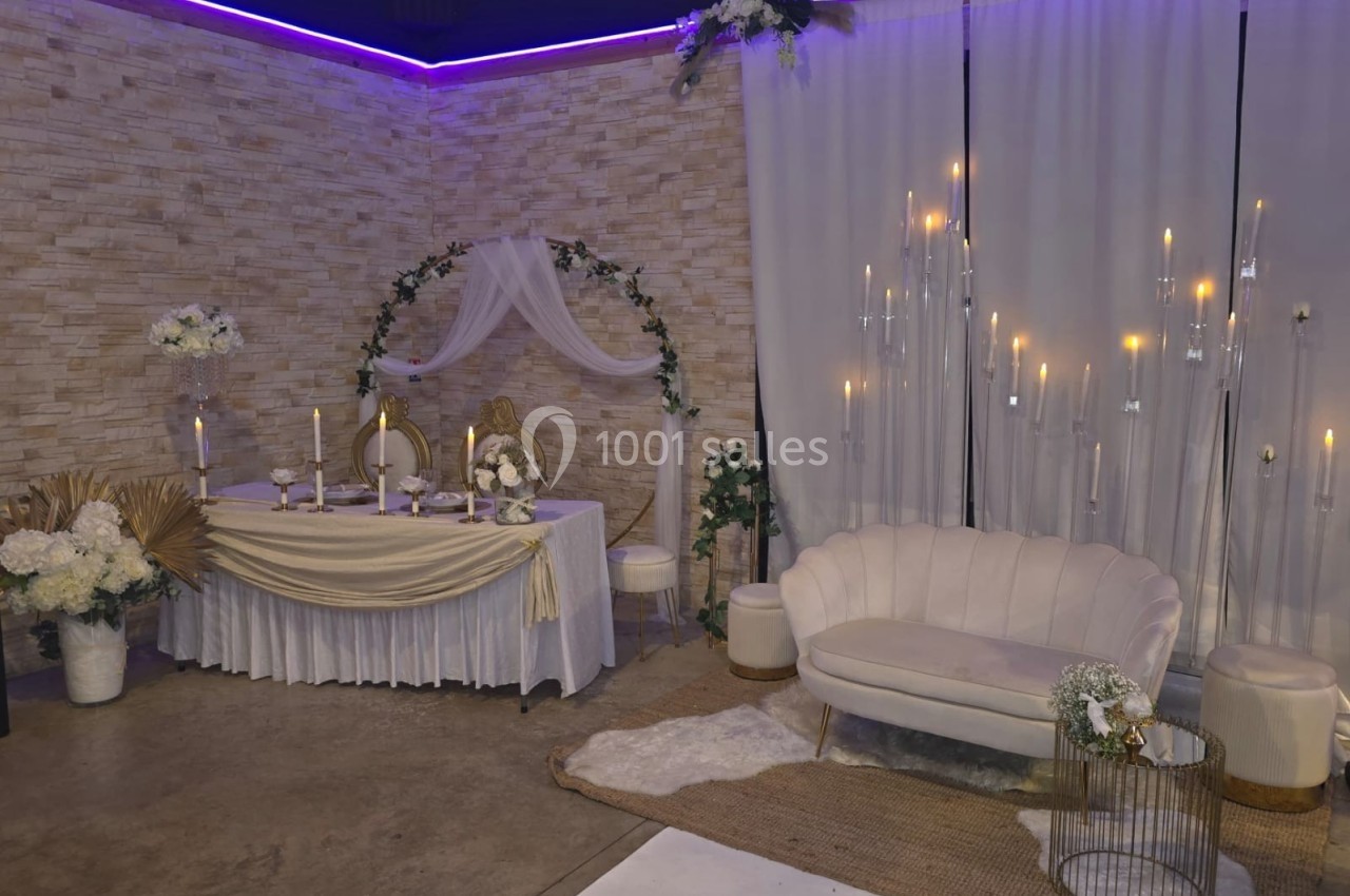 Décoration de salle avec table de réception ornée de fleurs, arche drapée, bougies et canapé blanc sur tapis beige.