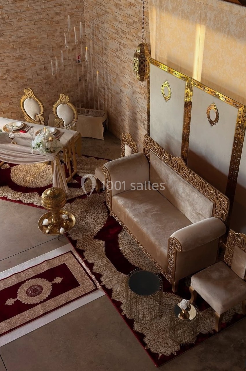 Salon décoré avec un canapé beige, tapis ornés, table avec chaises élégantes et éléments dorés sur fond de mur en pierre.