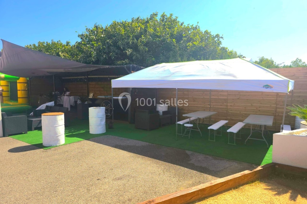Tonnelle blanche et espace ombragé avec tables, chaises et tonneaux, sur une terrasse extérieure ensoleillée.