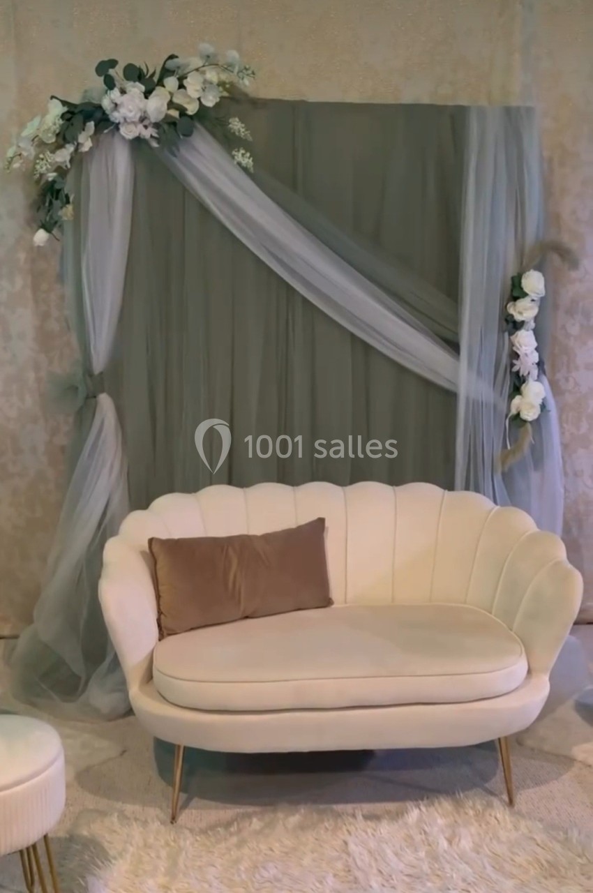 Canapé beige devant un fond décoratif avec rideaux en tulle gris et fleurs blanches.